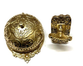 May include: A gold-toned doorbell with an ornate design. The doorbell has a round bell and a separate push button. The bell and button are both decorated with intricate floral patterns.