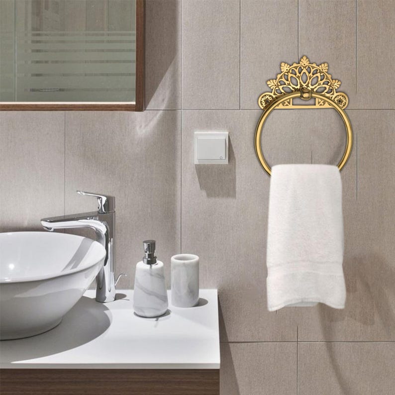 May include: A gold-colored towel ring with a decorative crown design is mounted on a tiled wall. A white towel hangs from the ring. The bathroom setting includes a sink, faucet, and soap dispenser.
