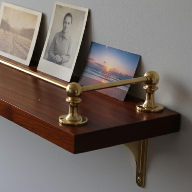 May include: A dark wood shelf with brass accents, holding three vintage photographs. The shelf has a brass rod and decorative supports. The photographs depict landscapes and portraits. The shelf is mounted on a light gray wall.