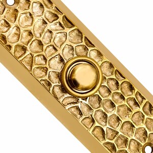 May include: A rectangular brass doorbell plate featuring a textured, hammered surface that resembles a pattern of small stones. The plate includes a centered, circular push button and two mounting holes at the top and bottom ends.