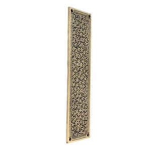 May include: A rectangular brass door plate with a detailed floral pattern. The plate has a dark background with a repeating light-colored floral design. The edges are lined with small dots, and there are four screw holes.