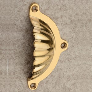 Puede incluir: Un tirador de armario de lat&oacute;n pulido con un dise&ntilde;o en forma de concha. El tirador tiene una forma curva con una serie de crestas verticales y tres orificios de montaje. El lat&oacute;n tiene una superficie brillante y reflectante.