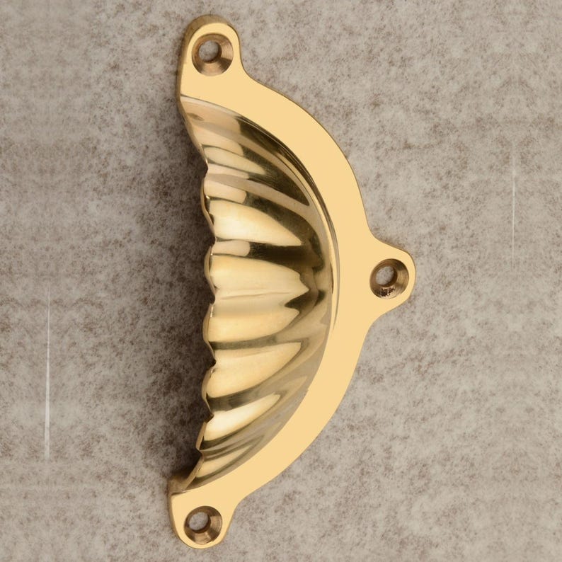 Puede incluir: Un tirador de armario de lat&oacute;n pulido con un dise&ntilde;o en forma de concha. El tirador tiene una forma curva con una serie de crestas verticales y tres orificios de montaje. El lat&oacute;n tiene una superficie brillante y reflectante.