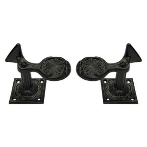 May include: Two black metal curtain rod holders. Each holder has a square base with a floral design, a vertical post, and a decorative top with an oval shape and leaf-like details. The holders are symmetrical and designed for mounting.