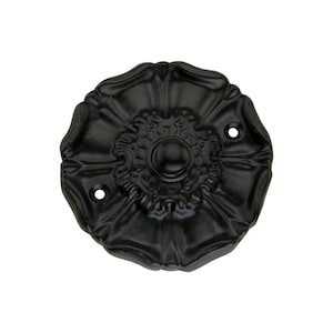 Pode incluir: Um botão de campainha preto e ornamentado com um design floral. O botão tem um elemento circular central e dois pequenos orifícios para montagem. A forma geral é redonda com um acabamento fosco texturizado.