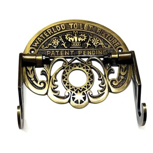 May include: Antique brass toilet paper holder with ornate scrollwork and a crown design. The holder has the text "Waterloo Toilet Fixture Patent Pending" on the top.