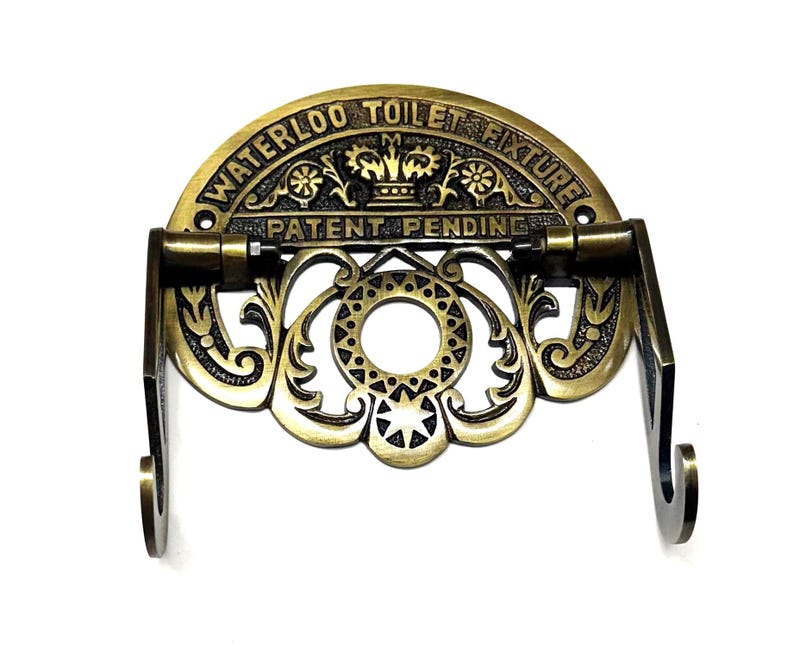 May include: Antique brass toilet paper holder with ornate scrollwork and a crown design. The holder has the text "Waterloo Toilet Fixture Patent Pending" on the top.
