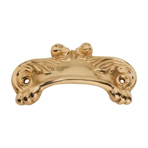 Op de afbeelding: Een goudkleurige, sierlijke kastgreep met een gebogen handvat. De greep heeft gedetailleerd krulwerk en twee decoratieve finials aan de bovenkant. Twee gaten zijn zichtbaar voor montage. De hardware heeft een gepolijste afwerking.
