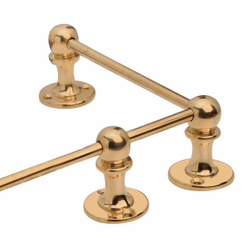 May include: A gold-colored towel rack with a classic design. The rack features three rounded supports and horizontal bars. The supports have a circular base with screw holes. The metal has a polished finish.