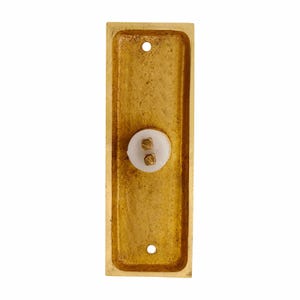 May include: A rectangular brass doorbell plate with a textured, metallic gold finish. The plate features a central white circular button component with two small brass screw terminals exposed, and two mounting holes at the top and bottom ends.