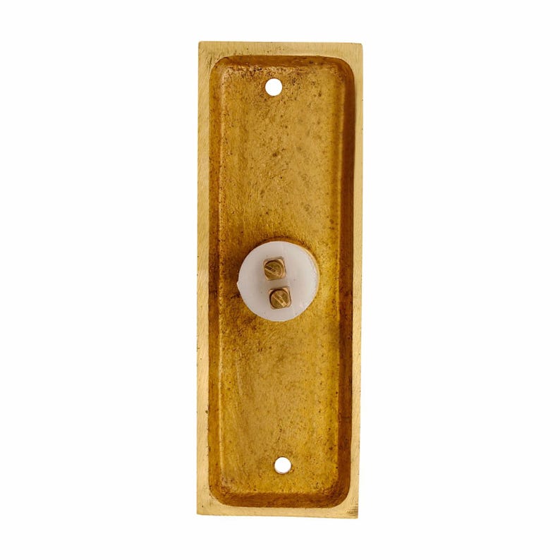 May include: A rectangular brass doorbell plate with a textured, metallic gold finish. The plate features a central white circular button component with two small brass screw terminals exposed, and two mounting holes at the top and bottom ends.