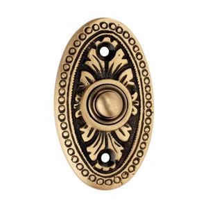 May include: Oval-shaped brass doorbell button with an ornate design. The button features a central circular push button, surrounded by a floral pattern and a border of small circles. Two screw holes are visible.