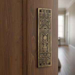 May include: A rectangular, antique brass door pull with an ornate floral design. The pull features detailed engravings of flowers, a vase, and geometric patterns. It is mounted on a wooden door, showcasing a classic, decorative style.