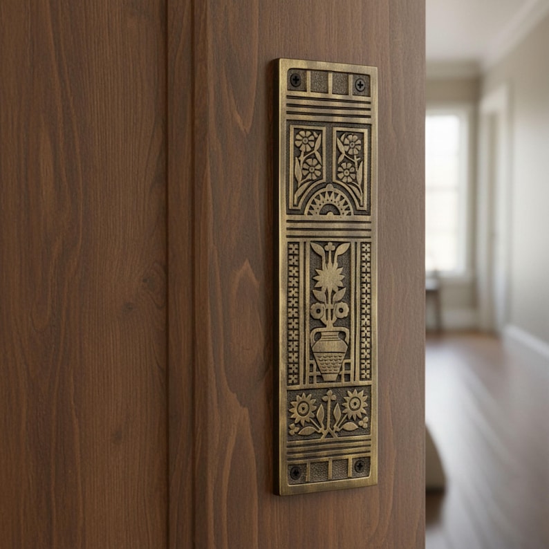 May include: A rectangular, antique brass door pull with an ornate floral design. The pull features detailed engravings of flowers, a vase, and geometric patterns. It is mounted on a wooden door, showcasing a classic, decorative style.