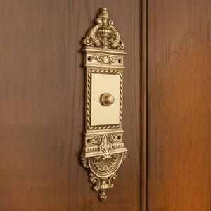 May include: An antique brass doorbell, featuring intricate scrollwork and a central button. The rectangular face is cream-coloured, framed by a rope-style border. It's mounted on a dark wooden door, adding a touch of elegance.