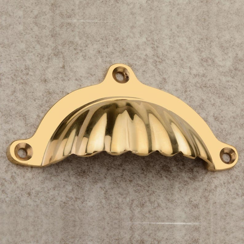 Puede incluir: Un tirador dorado en forma de concha con un interior acanalado. El herraje tiene tres orificios para tornillos para el montaje. El tirador est&aacute; hecho de un metal brillante y reflectante.