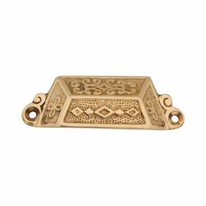 Puede incluir: Un tirador de cajón de metal dorado con forma trapezoidal y superficie texturizada. El panel frontal muestra una fila central de formas de diamante, mientras que la sección superior tiene volutas. Cada lado tiene un orificio para tornillo.