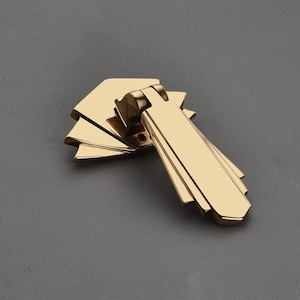 Op de afbeelding: Een goudkleurige kastgreep met een geometrisch ontwerp. De greep heeft een rechthoekige handgreep met gelaagde, waaiervormige details. De hardware is van metaal en heeft een glanzende afwerking.