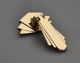 May include: A gold-colored cabinet pull with a geometric design. The pull features a rectangular handle with layered, fan-shaped details. The hardware is made of metal and has a shiny finish.