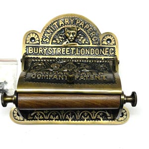 May include: Antique brass toilet roll holder with a wooden roller and ornate detailing. The holder features the text "SANITARY PAPER BURY STREET LONDON EC COMPANY PATENT".