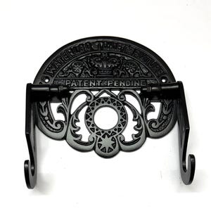 May include: Black cast iron toilet paper holder with a decorative design and the text "Waterloo Toilet Fixture Patent Pending".