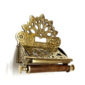 May include: A gold-toned brass toilet paper holder with an ornate, decorative design. The holder has a wooden roller and a shelf for extra toilet paper.