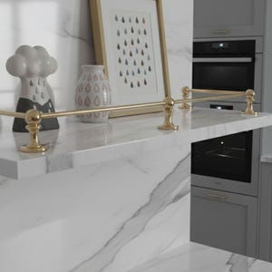 May include: A modern kitchen shelf with a gold-colored railing. The shelf is made of white marble and features decorative items, including a cloud-shaped sculpture and a vase. A framed picture with a teardrop design is also on the shelf.