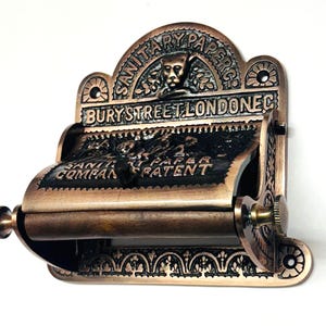 May include: Antique brass toilet paper holder with intricate detailing. The holder features the text "SANITARY PAPER CO. BURY STREET LONDON EC."