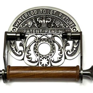 May include: Silver toilet roll holder with an ornate design and the words "Waterloo Toilet Fixture Patent Pending". The holder has a wooden roller.