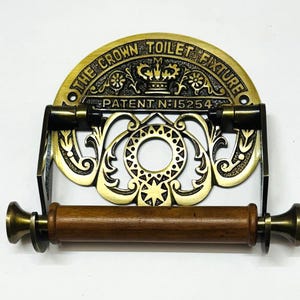May include: Antique brass toilet roll holder with a decorative scroll design and a wooden roller. The holder features a crown and the text "The Crown Toilet Fixture Patent N:15254".