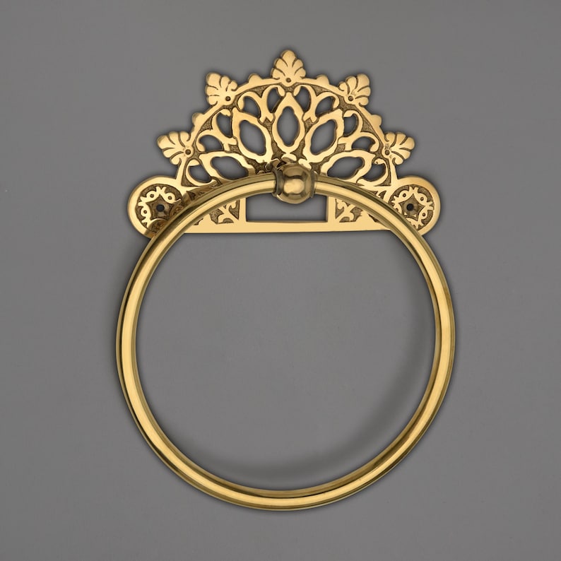 May include: A gold-colored towel ring with an ornate, decorative backplate. The backplate features a floral design with a semi-circular shape. The ring is circular and attached to the backplate with a small ball joint. The overall design is vintage.