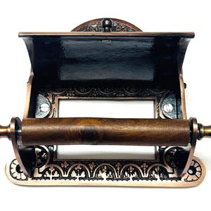 May include: Antique copper-toned toilet paper holder with an ornate design and a wooden roller. The holder has a built-in shelf for storing extra toilet paper.