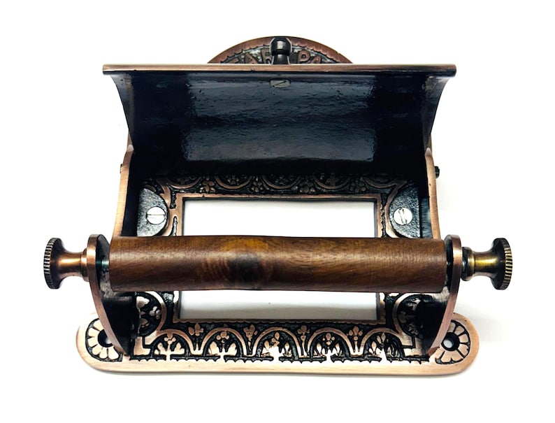 May include: Antique copper-toned toilet paper holder with an ornate design and a wooden roller. The holder has a built-in shelf for storing extra toilet paper.
