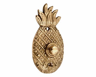 May include: A brass doorbell button shaped like a pineapple with a textured cross-hatched surface and a central circular push mechanism. The pineapple features a leafy top and two mounting holes at the top and bottom for installation on a wall.