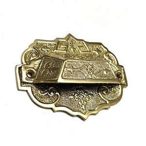 May include: A gold-toned metal door knocker with an ornate design. The knocker is rectangular in shape with a raised, decorative centre section. The edges of the knocker are scalloped and have a detailed floral pattern.