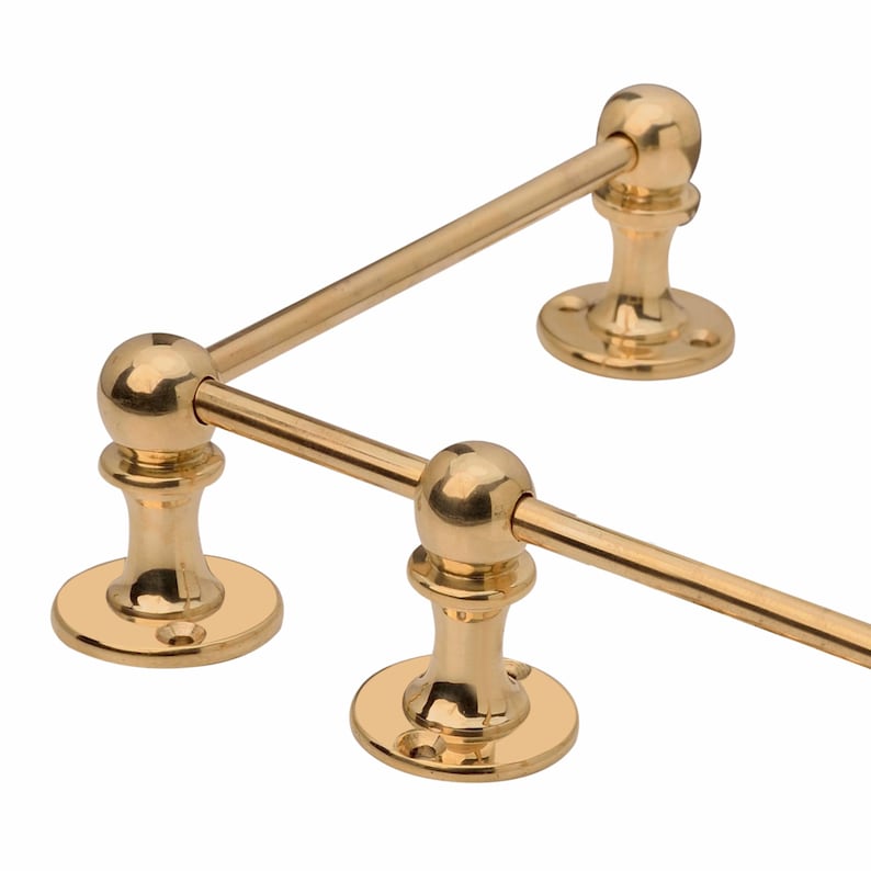 May include: A gold-colored, two-bar towel rack. The rack has a classic design with a round base and ball finials. The bars are horizontal and connected to the base with decorative supports. The rack is made of metal and has a polished finish.