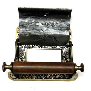 May include: A vintage brass toilet roll holder with an ornate design and a wooden roller. The holder has a black metal flap that can be lifted to access the roll.