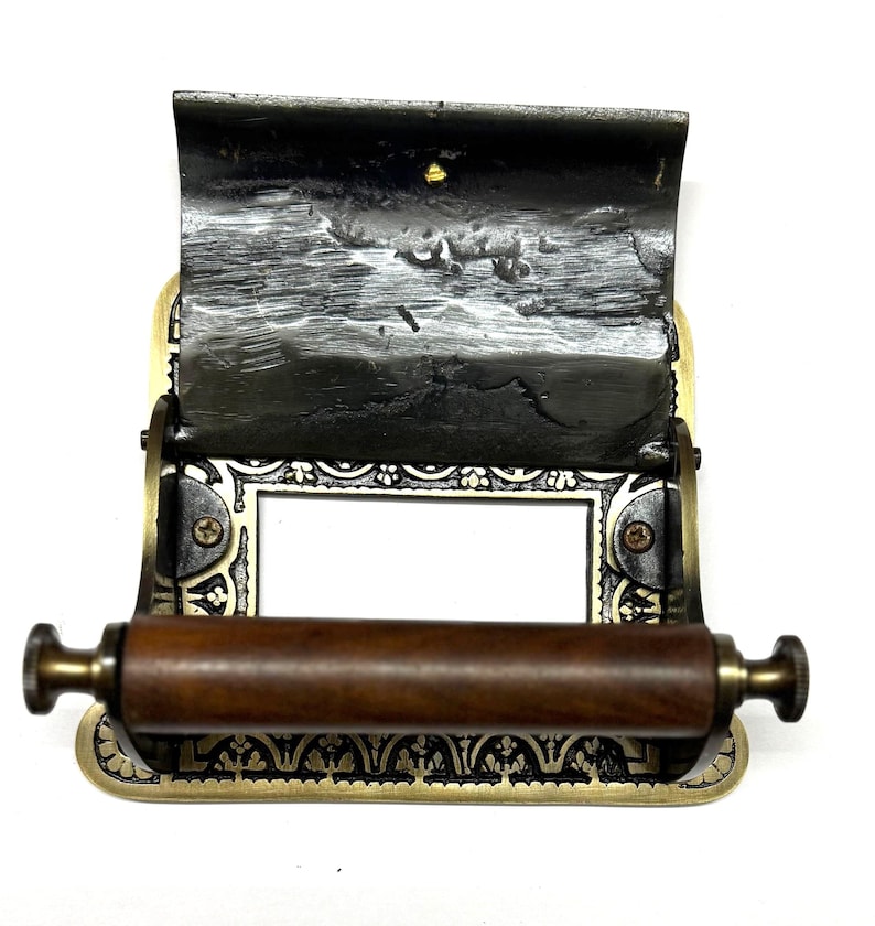 May include: A vintage brass toilet roll holder with an ornate design and a wooden roller. The holder has a black metal flap that can be lifted to access the roll.