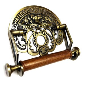 May include: Antique brass toilet paper holder with a decorative design and the text "Waterloo Toilet Fixture Patent Pending". The holder has a wooden roller.
