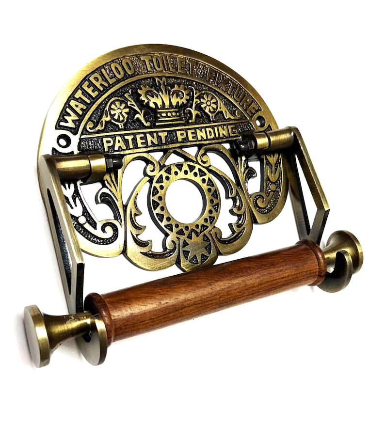 May include: Antique brass toilet paper holder with a decorative design and the text "Waterloo Toilet Fixture Patent Pending". The holder has a wooden roller.