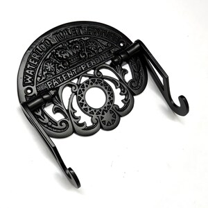 May include: Black cast iron toilet paper holder with ornate detailing and the text "Waterloo Toilet Fixture Patent Pending And".