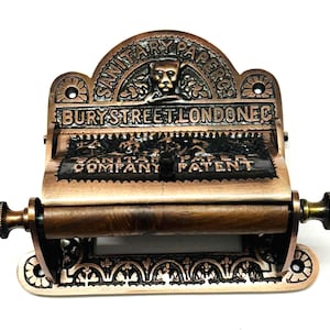 May include: Antique copper-toned toilet paper holder with intricate detailing. The holder features a decorative top with the text "SANITARY PAPERS CO. BURY STREET LONDON EC."