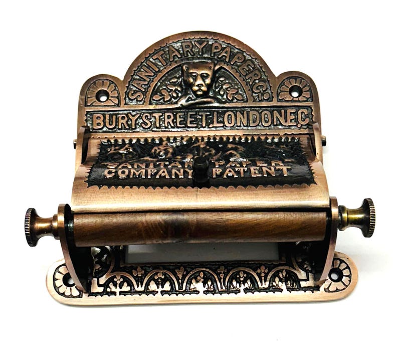 May include: Antique copper-toned toilet paper holder with intricate detailing. The holder features a decorative top with the text "SANITARY PAPERS CO. BURY STREET LONDON EC."