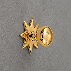 May include: A polished gold-tone door knob featuring a spherical handle attached to a decorative backplate shaped like an eight-pointed star. The metallic surface is reflective and smooth, showcasing a bright, reflective finish throughout.