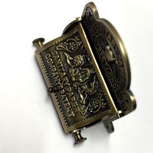 May include: Antique brass toilet paper holder with the text "SANITARY PAPER COMPANY'S PATENT" and "ALPHABURY STREET LONDON EC" embossed on the front.