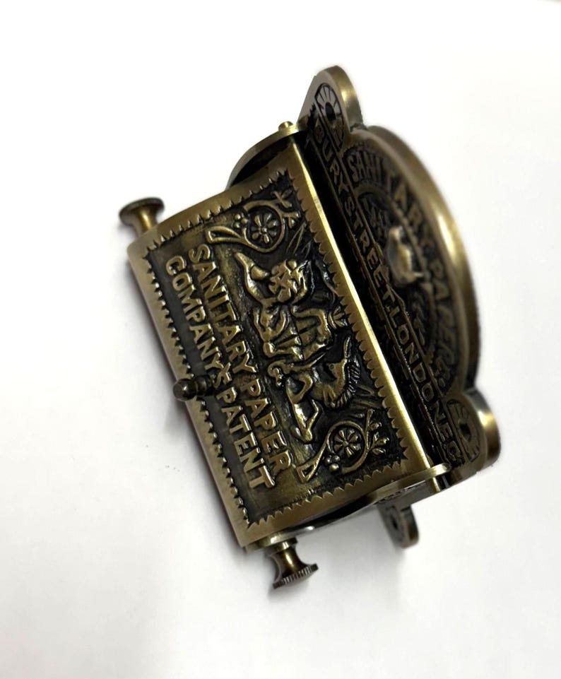 May include: Antique brass toilet paper holder with the text "SANITARY PAPER COMPANY'S PATENT" and "ALPHABURY STREET LONDON EC" embossed on the front.