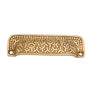 May include: A gold-coloured, ornate drawer pull with a floral pattern. The rectangular pull has a curved handle and two holes for mounting. The design features a detailed, embossed floral motif and a dotted border.