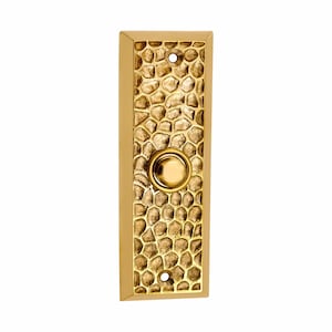 May include: A rectangular brass doorbell plate featuring a textured, hammered surface pattern. The plate has a smooth, beveled border and a centered circular push button. Two small screw holes are visible at the top and bottom of the plate.
