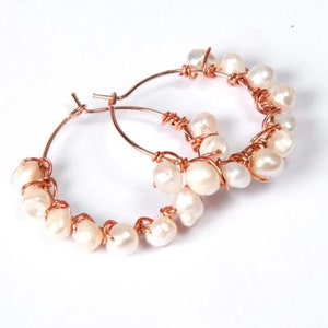 以下が含まれることがあります： ローズゴールドのフープピアス。白い淡水パールがフープに巻き付けられています。