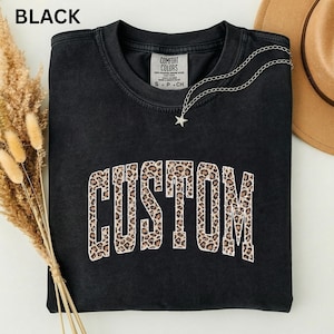 Könnte beinhalten: Ein schwarzes Sweatshirt mit Rundhalsausschnitt und dem Wort CUSTOM in einer großen, geschwungenen Schrift mit Leopardenmuster und weißer Umrandung. Das Kleidungsstück hat ein Comfort Colors-Etikett am Halsausschnitt und eine silberne Sternkette.